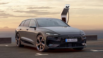 Der A6 Avant e-tron steht rückwärts auf einem Parkplatz und ist dabei an eine Ladestation angeschlossen. Im Hintergrund sind das Meer, sowie Berge zu erkennen.