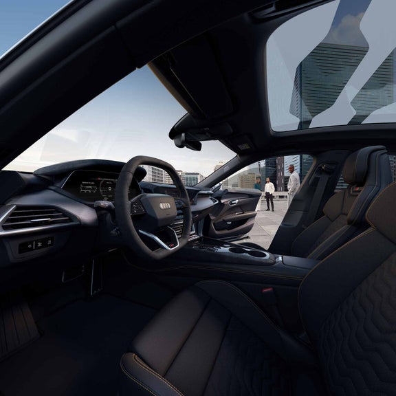 Innenraum des Audi S e-tron GT mit sportlichem Lenkrad, digitalen Anzeigen und hochwertigen Materialien. Durch das offene Fenster sind drei Personen auf einer Dachterrasse mit Stadtblick zu sehen.