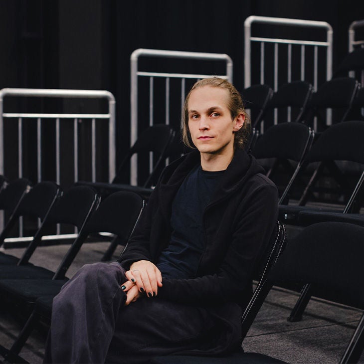 Junger Mann mit langen blonden Haaren sitzt auf schwarzen Stühlen in einem leeren Saal.