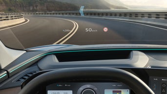 Detailaufnahme des Innenraum-Cockpits eines Audi, frontal von Fahrerseite aufgenommen. Männerhände greifen das Lenkrad. Auf die Windschutzscheibe sind Verkehrszeichen projiziert.