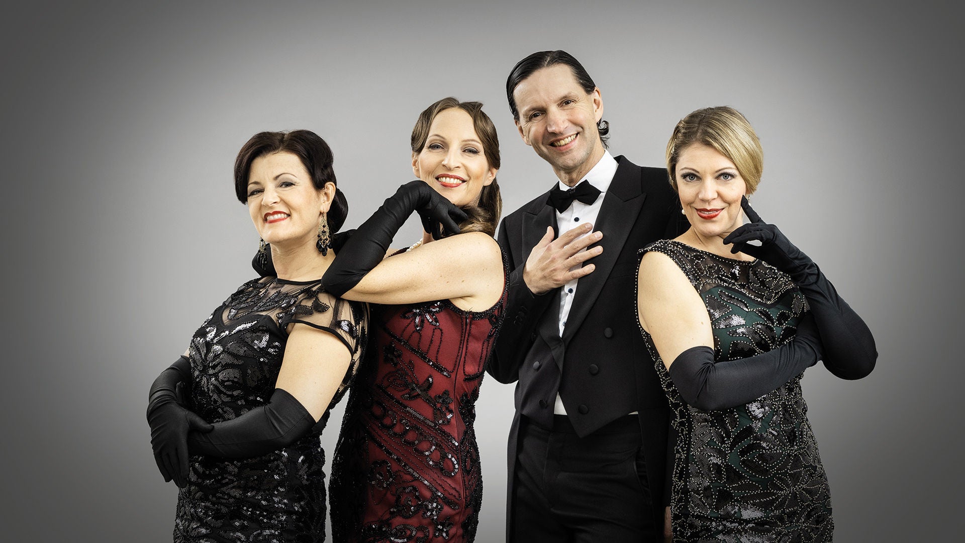 Drei Frauen in eleganten Kleidern und ein Mann im Anzug posieren lächelnd vor grauem Hintergrund.