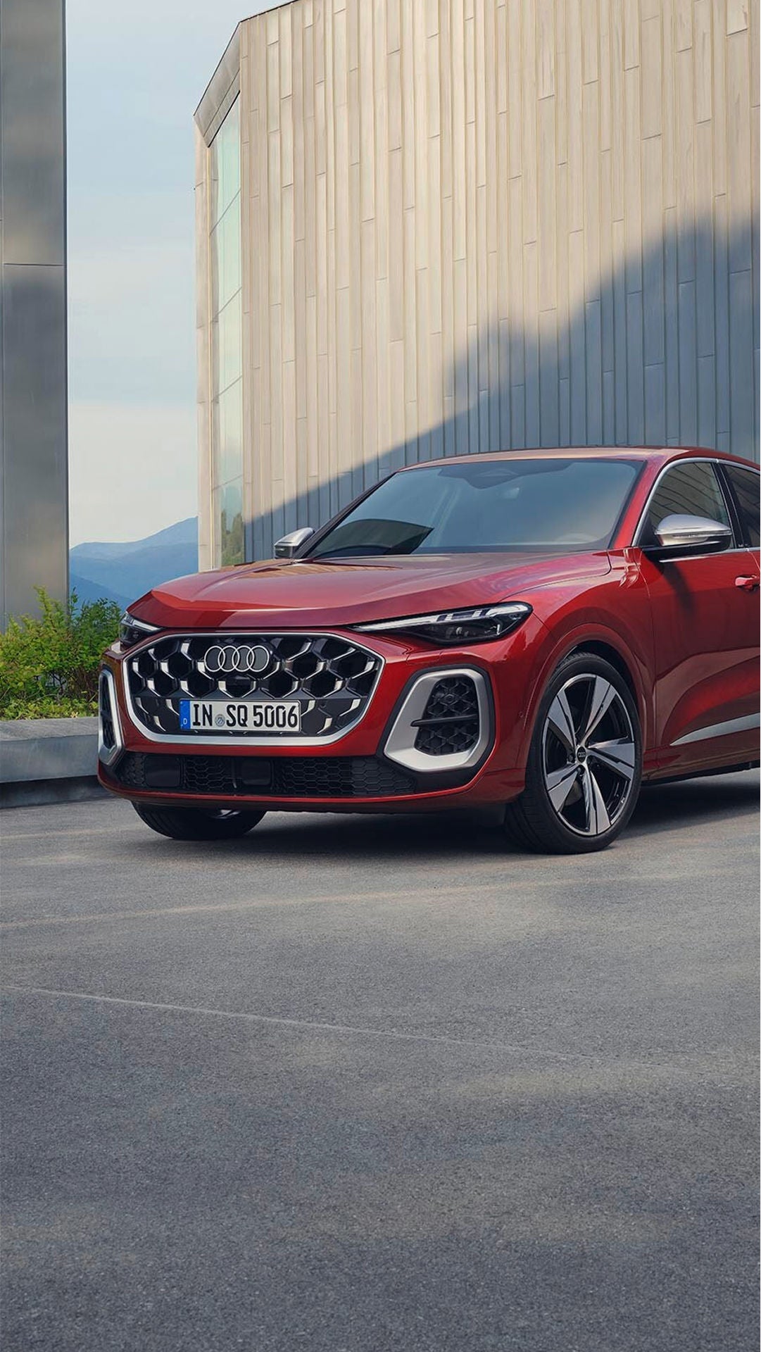 Red Audi parked on a modern concrete driveway, featuring sleek lines and large alloy wheels. The backdrop includes green foliage and a geometric building facade, conveying a sense of elegance and luxury.