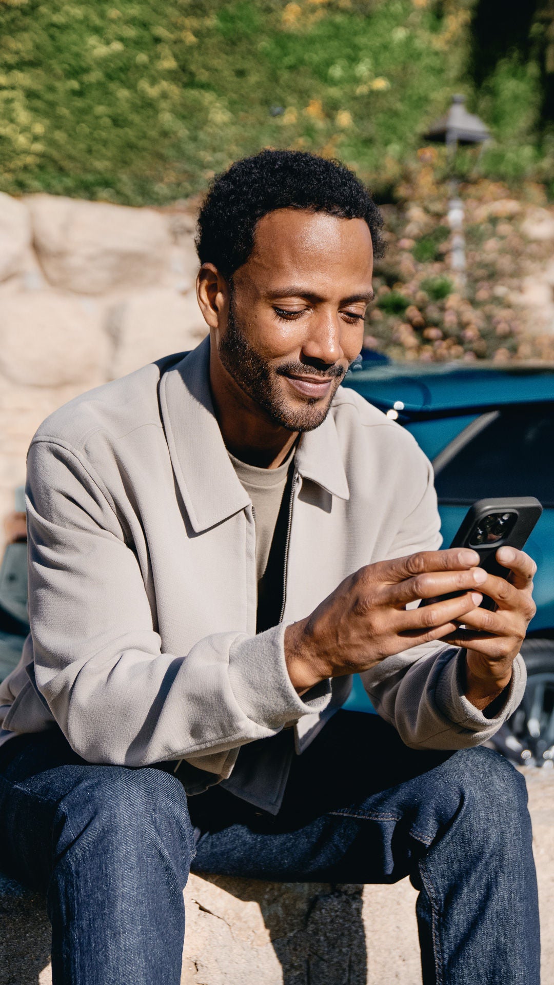 Eine Frau in rotem Hosenanzug sitzt im Freien an einem Laptop. Im Hintergrund steht ein blaues Fahrzeug vor einem Gebäude.