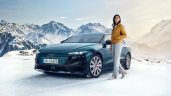 Eine Frau in einer gelben Winterjacke steht neben einem petrolfarbenen Audi S6 e-tron in einer Winterlandschaft. Im Hintergrund sind schneebedeckte Gipfel zu sehen.