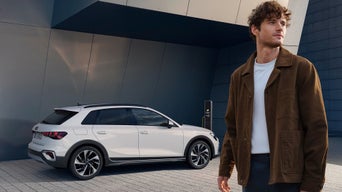 Ein Mann steht im Vordergrund und schaut nach rechts, während ein weißer Audi A3 hinter ihm an einer Ladestation steht