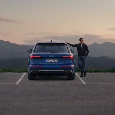 Ein blauer Audi Q7 SUV von hinten, asiatischer Mann lehnt sich mit Hand rechts am Fahrzeug an, Berge bei Sonnenuntergang im Hintergrund.