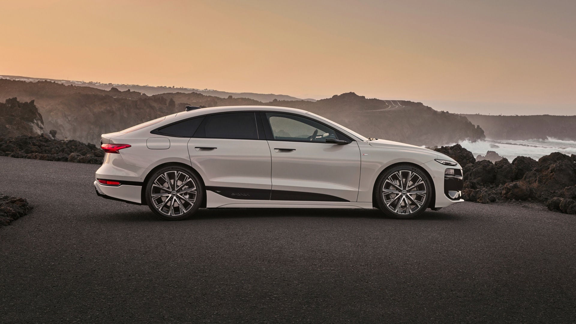 A sleek white luxury sedan parked on a coastal road, against a backdrop of rocky cliffs and a pastel sunset sky.
