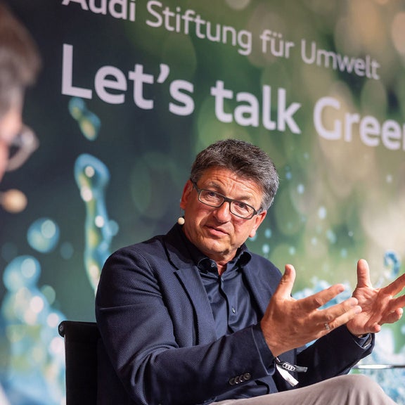 A man in a jacket speaks on a stage and gesticulates while a presenter listens to him. The background shows “Let's talk Greenovation”.