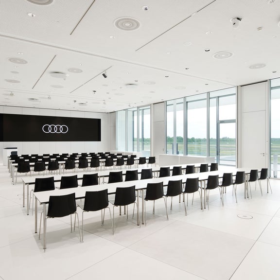 Heller Konferenzraum mit bodentiefen Fenstern, schwarzer Display mit Audi Logo, weißen Tischen und schwarzer Bestuhlung. 