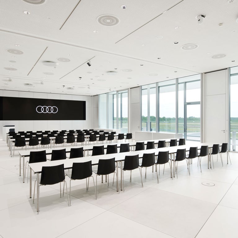 Heller Konferenzraum mit bodentiefen Fenstern, schwarzer Display mit Audi Logo, weißen Tischen und schwarzer Bestuhlung. 