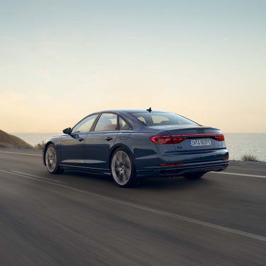 Ein dunkelblauer Audi A8 60 TFSI e fährt bei Sonnenuntergang auf einer Straße am Meer entlang, Rückansicht.