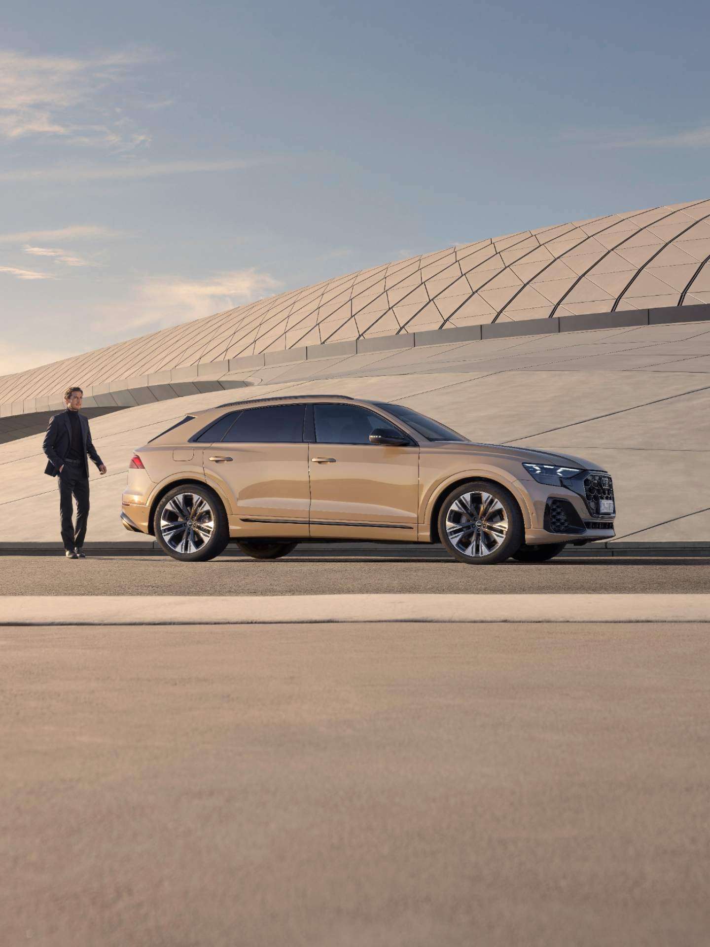 Man standing behind beige coloured Audi Q8 SUV with modern architecture in background.