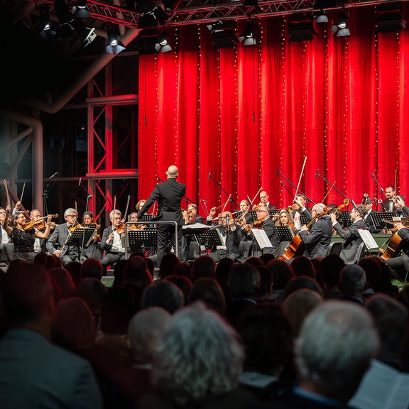 Dirigent leitet großes Orchester vor rotem Vorhang, Publikum im Vordergrund; feierliche Konzertszene mit klassischer Musik.