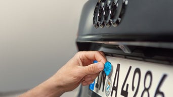 Hand bringt blaue Prüfplakette am Kennzeichen eines Audi-Fahrzeugs an.