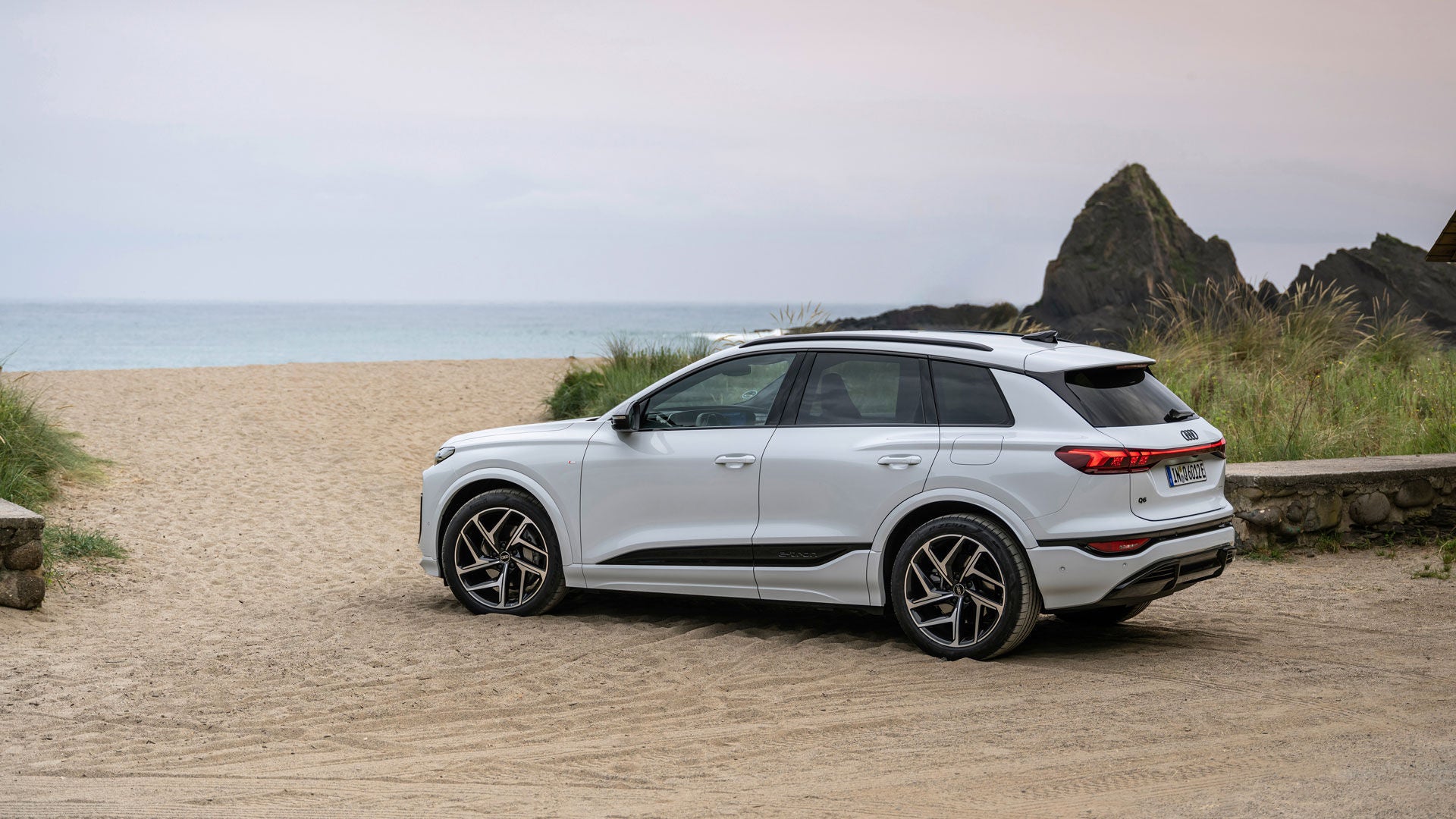 Weißer Audi Q6 SUV e-tron an einem Strandübergang. Hinter dem Fahrzeug ist ein breiter Sandstrand vor dem offenen Meer.