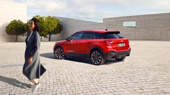 Roter Audi SQ2 auf Kopfsteinpflaster, lächelnde Frau in grauem, langen Mantel geht im Vordergrund, moderne Architektur im Hintergrund.