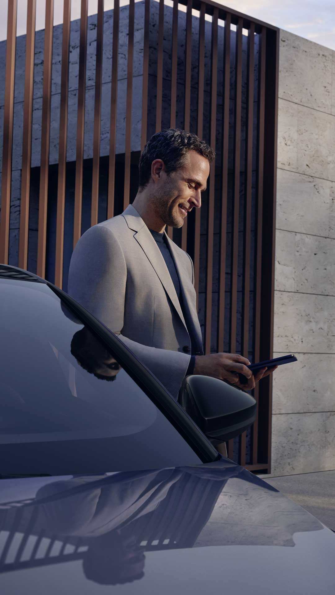 Mann mit Smartphone vor weißem Audi, nutzt Fahrzeug-App; im Hintergrund modernes Holzgebäude und grüne Rasenfläche.