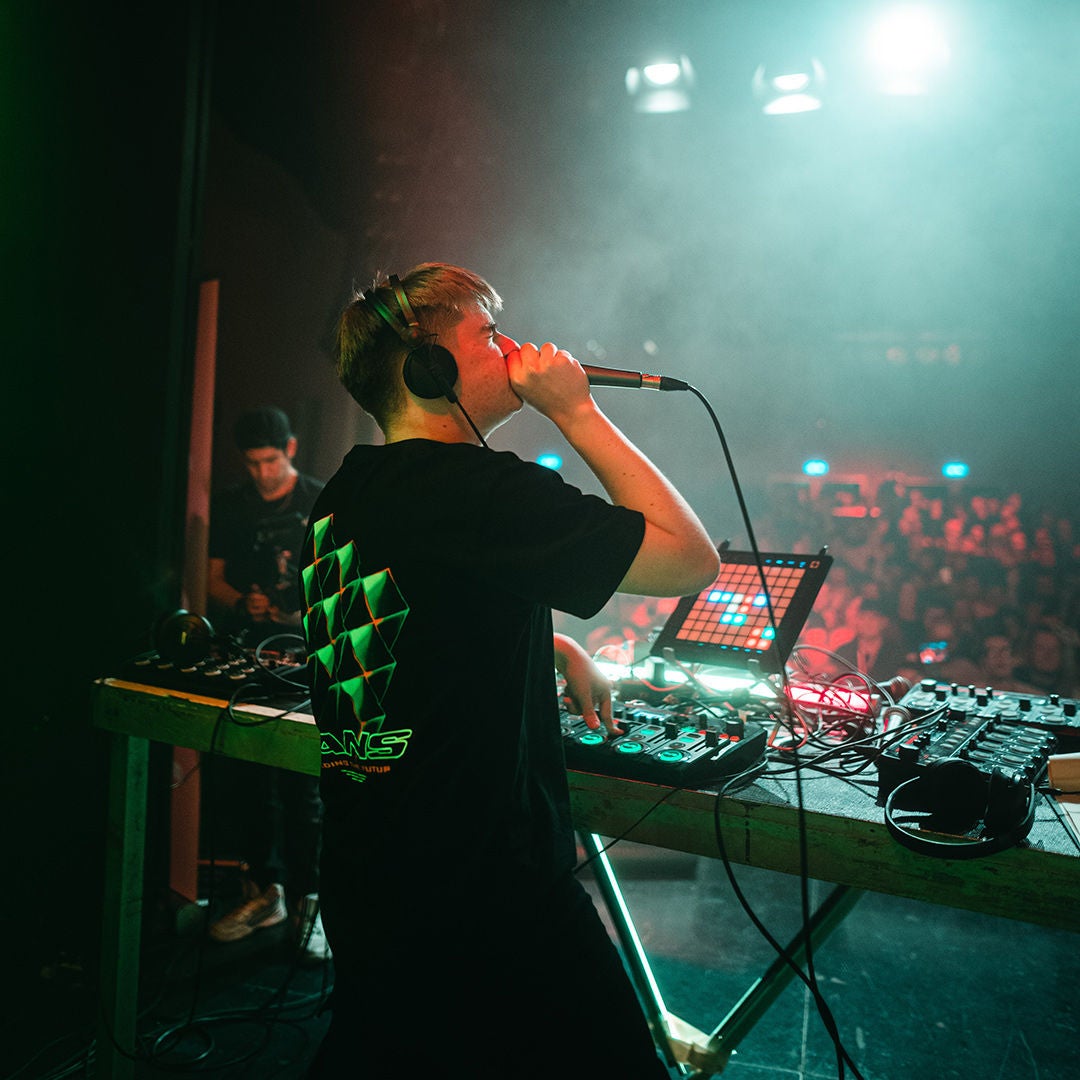 DJ/Live-Act mit Kopfhörern am Mischpult, singt ins Mikrofon; grün-rotes Bühnenlicht und Publikum im Hintergrund.