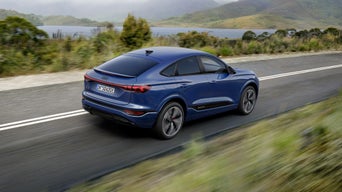 Seitliche Heckaufnahme des SQ6 Sportback e-tron in dunkelblau, welcher eine Straße in der Natur neben einem See entlangfährt.