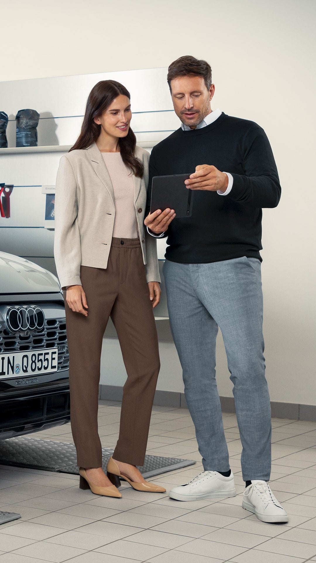 Mann zeigt Frau etwas auf einem Tablet neben einem grauen Audi in einer Werkstatt mit Felgen und Zubehör an der Wand.