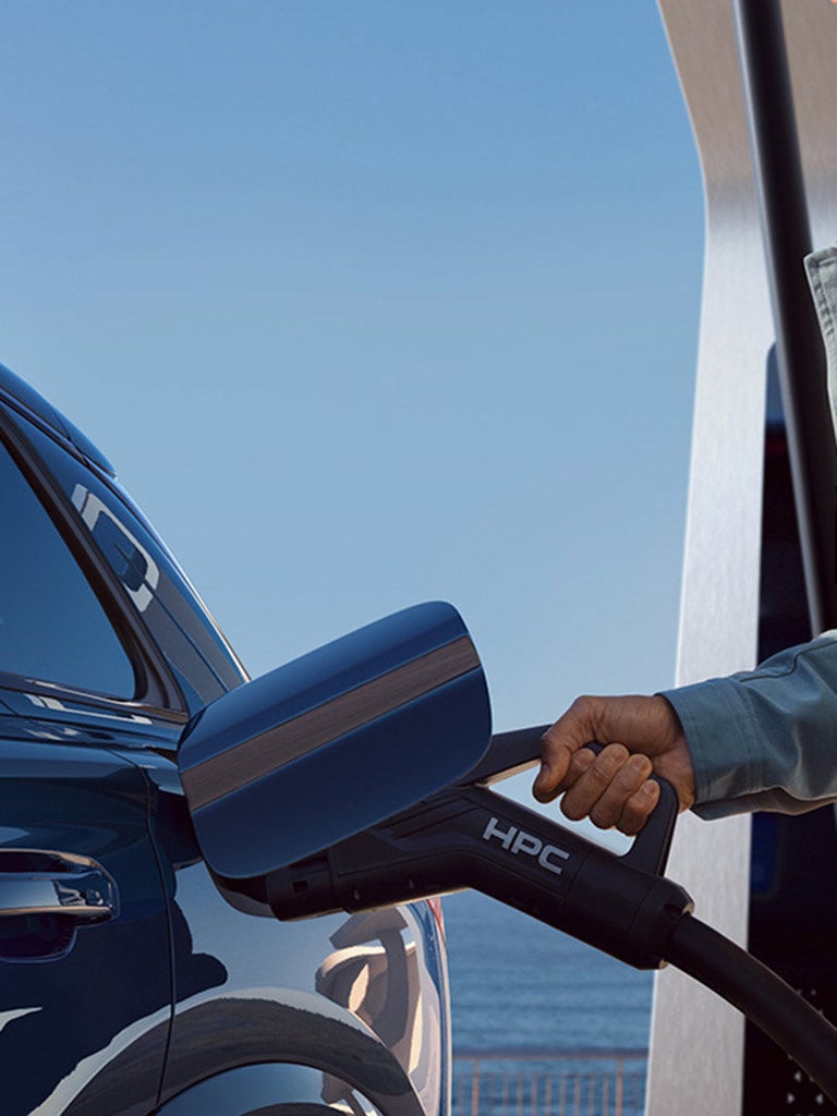 Mann lädt blauen Audi Q6 SUV e-tron an einer Ladesäule mit Blick auf das Meer im Hintergrund.