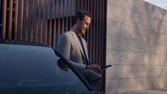 Ein lächelnder, grau gekleideter Mann steht vor einem futuristischen Gebäude und blickt auf ein Smartphone. Sein Unterkörper wird von einem Audi verdeckt, dessen Windschutzscheibe von links ins Bild ragt.