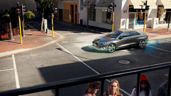 Ein grauer Audi fährt über städtische Kreuzung bei Tageslicht (digitales Linien zeigen Assistenzsysteme), Passantinnen stehen im Vordergrund am Straßenrand.