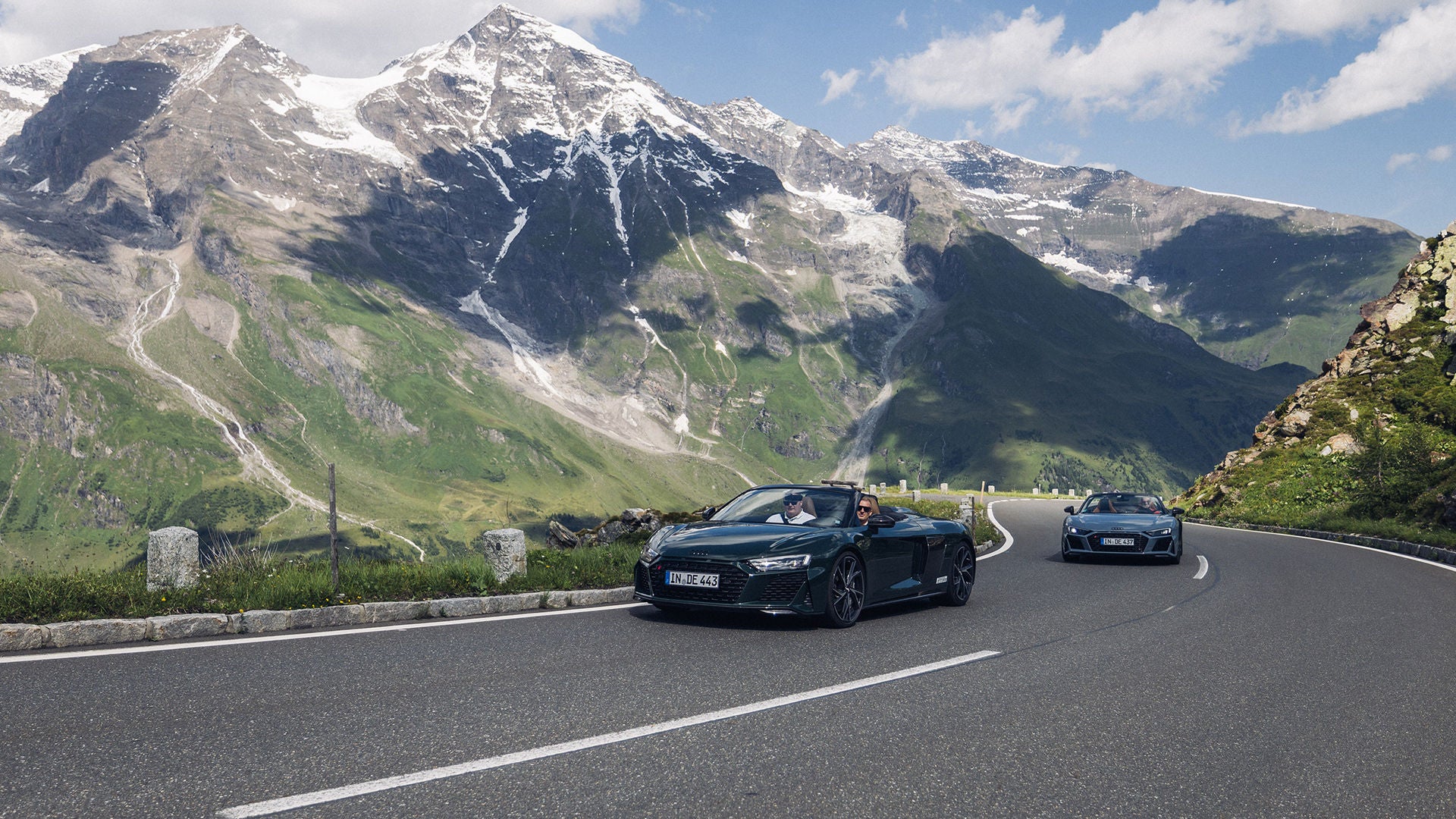 Two Audis drive along a winding mountain road in front of snow-covered peaks in sunny weather.