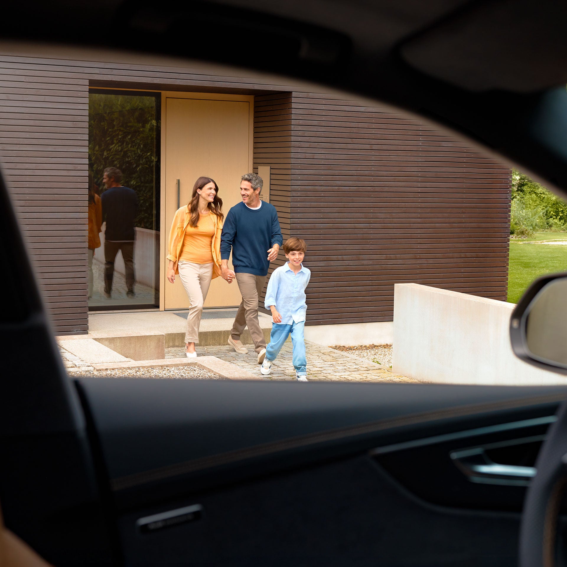 Blick aus einem Auto auf eine Familie mit Kind vor einem Haus auf dem Weg zum Fahrzeug.