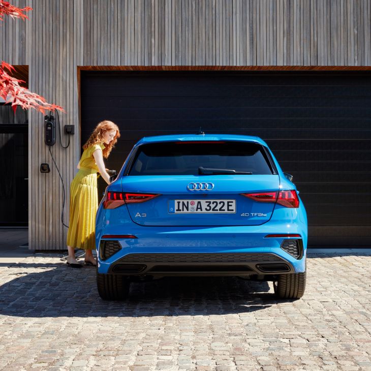 A blue Audi A3 parked in front of a modern garage with a person in a yellow dress beside it.