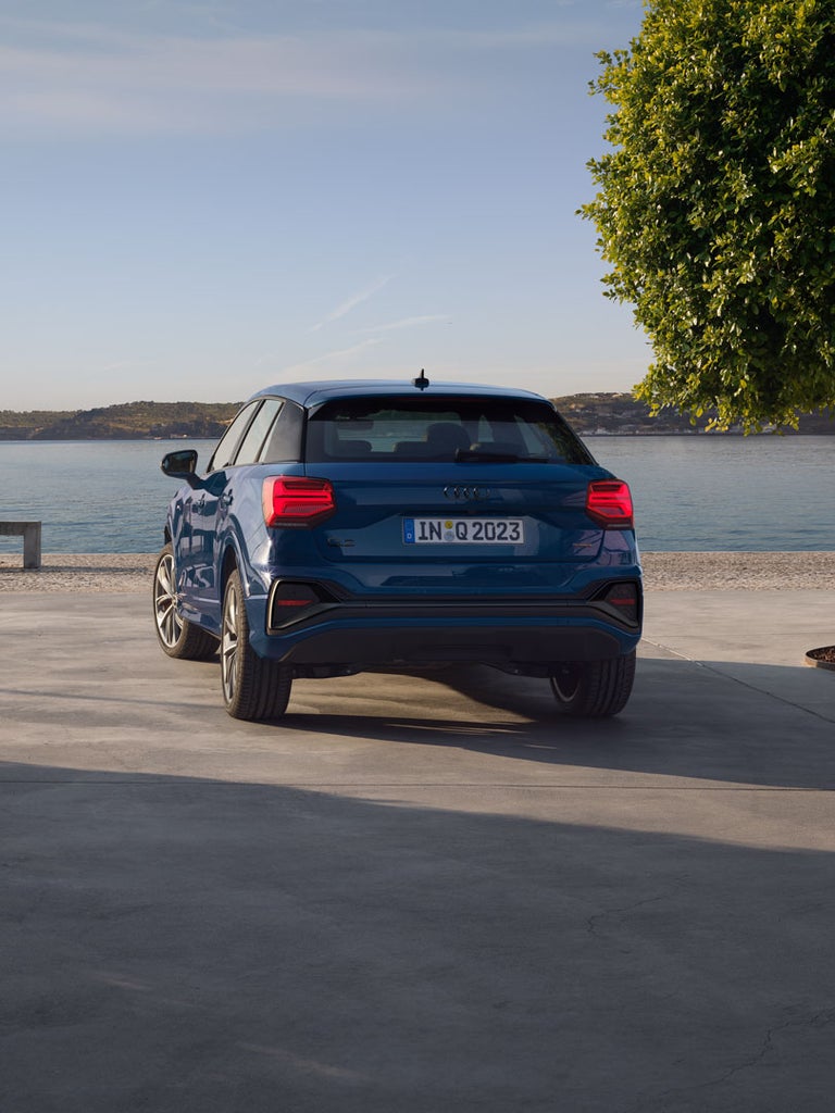 Ein blauer Audi Q2 von hinten auf Platz am Wasser, Mann in weißem Pullover sitzt auf einer Betonbank am Ufer.