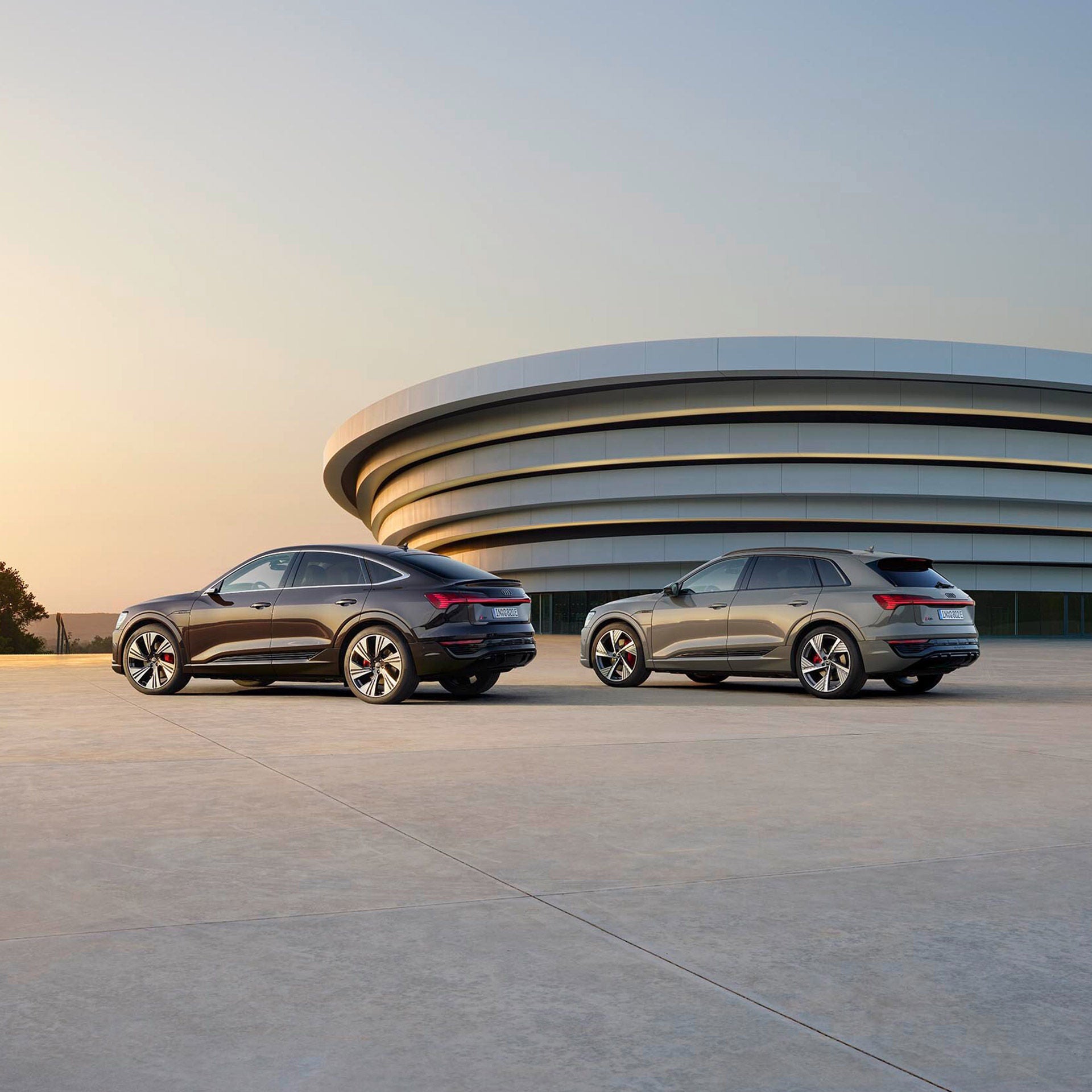 Zwei Elektrofahrzeuge von Audi vor einem modernen, runden Gebäude bei Sonnenuntergang auf einer offenen Betonflächen.