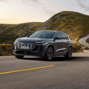 A grey Audi SUV drives on a winding road through scenic, rolling hills under a soft, overcast sky. The vehicle exudes modern elegance and adventure.
