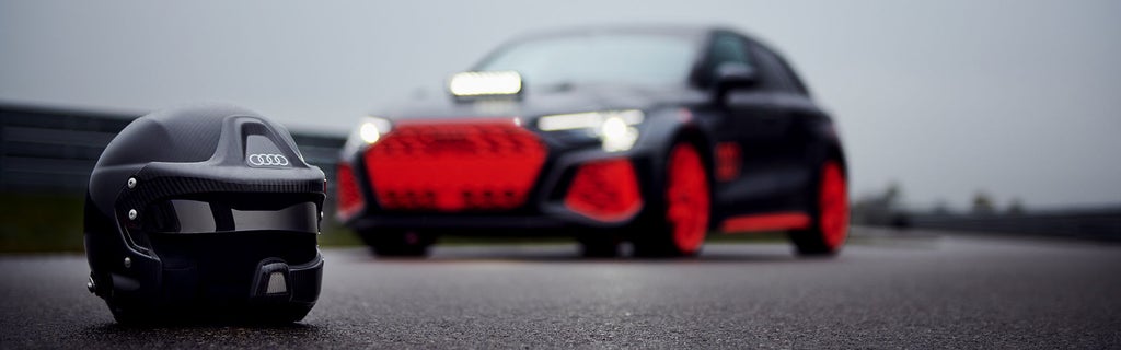 Schwarzer Rennhelm mit weißem Audi Logo auf nasser Rennstrecke, im Hintergrund unscharfer mattgrauer Audi RS 3 driving experience mit roten Akzenten bei grauem Himmel.