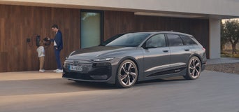 Grauer Audi A6 e-tron wird vor modernem Haus geladen; im Hintergrund stehen ein Mann und ein Kind an der Ladestation.