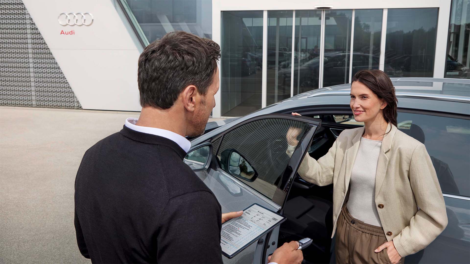 Eine Frau steigt vor einem Audi-Gebäude aus dem Auto, während ein Mitarbeiter mit Klemmbrett sie freundlich begrüßt.