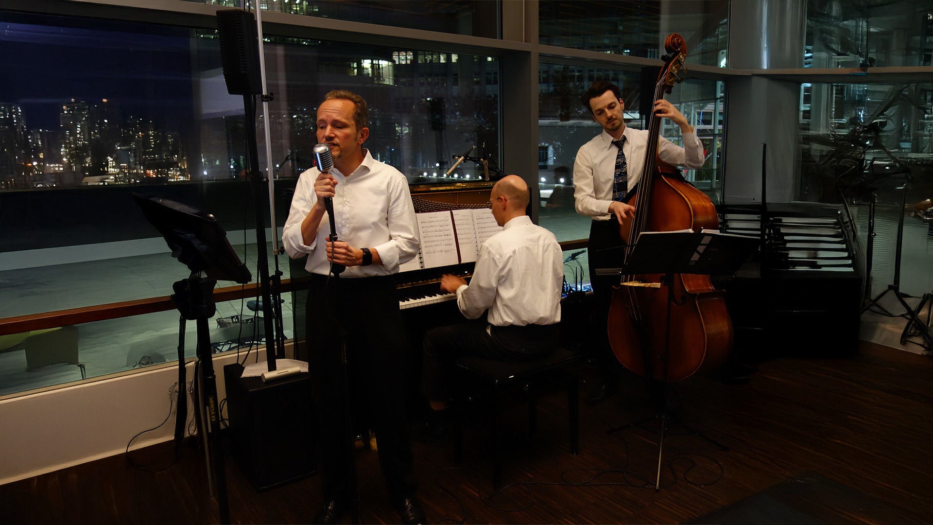 Jazztrio bei Abendveranstaltung: Sänger am Mikrofon, Pianist am Flügel und Kontrabassist spielen vor großer Glasfront bei Nacht.