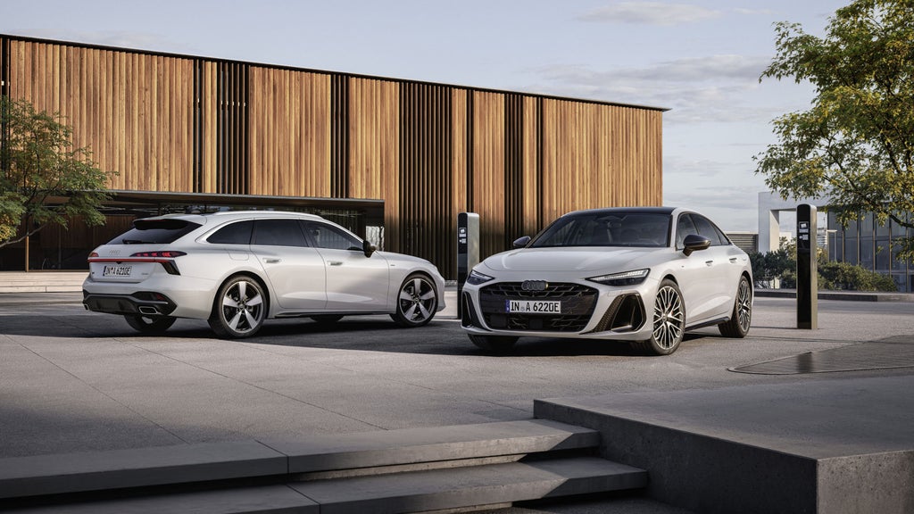 Ein weißer Audi A6 Avant e-hybrid quattro und eine weiße Audi A6 Limousine e-hybrid quattro stehen vor einem modernen Gebäude und sind an Ladestationen für Elektrofahrzeuge angeschlossen.