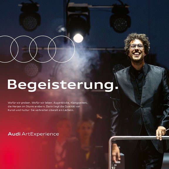 Dirigent mit Brille und Taktstock auf beleuchteter Bühne, lächelnd vor dunklem Hintergrund, Text „Begeisterung“.