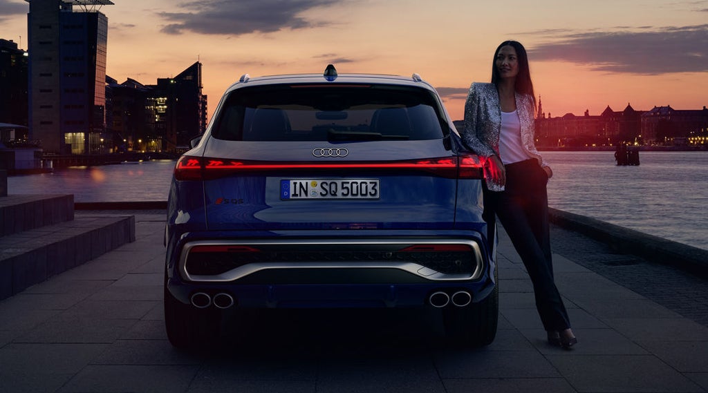 Das Heckprofil des SQ5 SUV steht vor der Skyline einer Küstenstadt. An das Heck lehnt eine junge Frau.