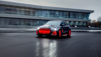 Audi RS 3 driving experience driving on a practice track under a gray sky at dusk, with a large building with glass fronts in the background.