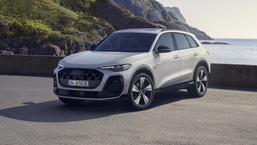 Der Q5 SUV in der edition one mit Kontrasten in Mythosschwarz steht im seitlichen Frontprofil auf einem Parkplatz direkt an der Küste. Dahinter ein futuristisches Haus.