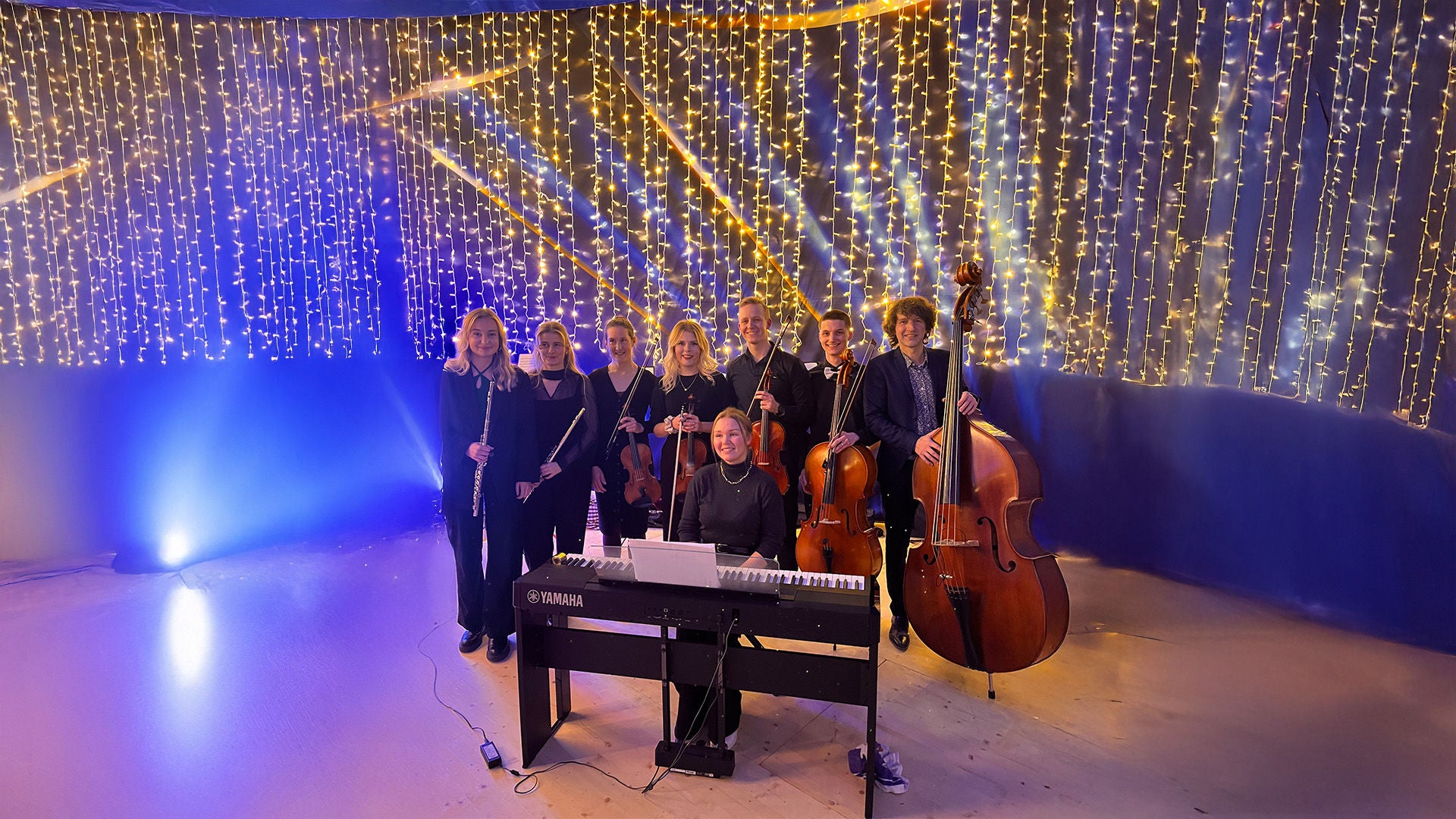 Musikensemble mit Streich- und Blasinstrumenten steht um ein E-Piano vor leuchtender Lichterkette.