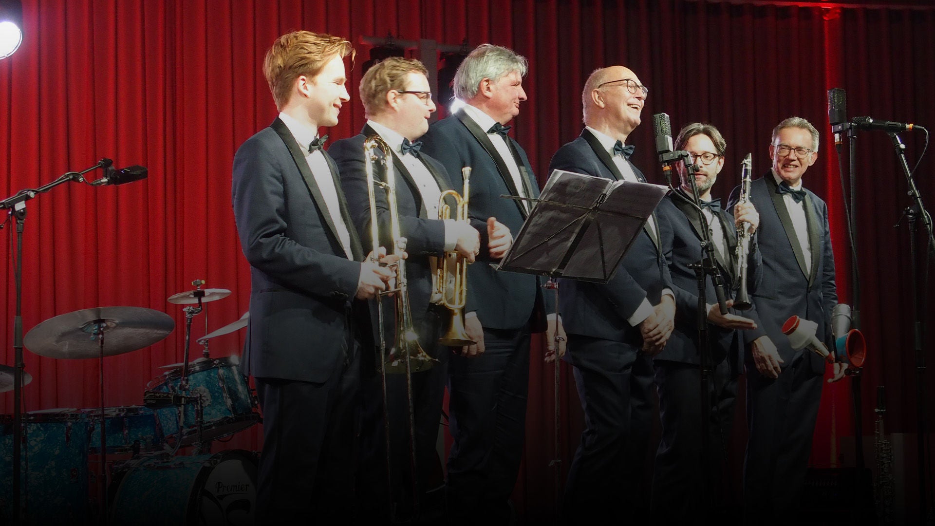 Jazzband in eleganten Smokings auf Bühne vor rotem Vorhang; mehrere Blechbläser und Mikrofone, daneben ein Schlagzeug im Hintergrund.