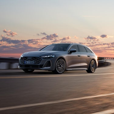 Grauer Audi S5 Avant fährt auf kurviger Küstenstraße bei Sonnenuntergang, von schräg vorn gezeigt.