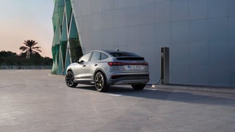 Silberner Audi Q4 e-tron lädt an einer Säule vor einem modernen Gebäude mit Glasfassade und Palmen im Hintergrund.