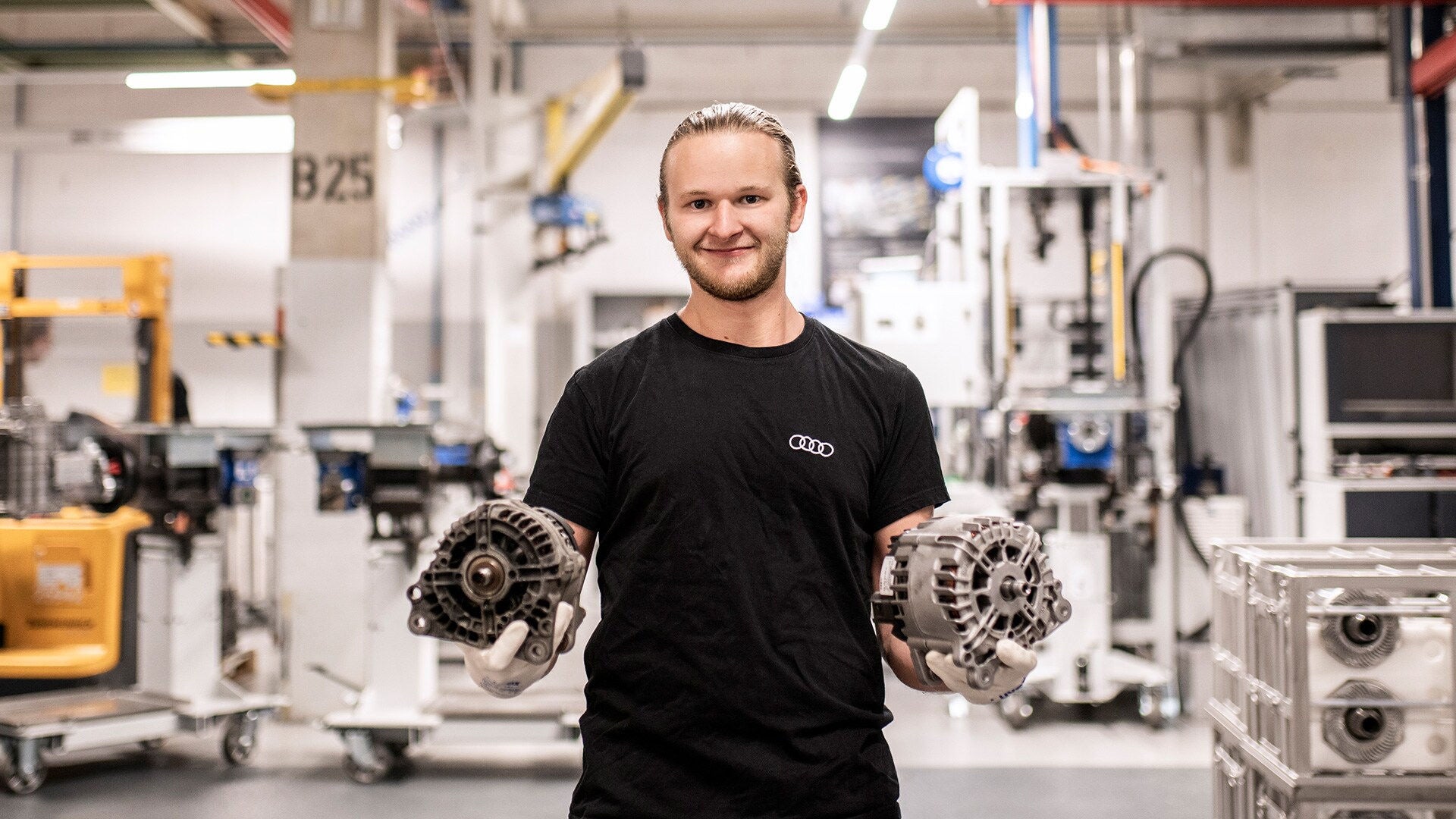 A person wearing a t-shirt with the Audi rings holds two metal components in an industrial production environment with machines and workbenches.