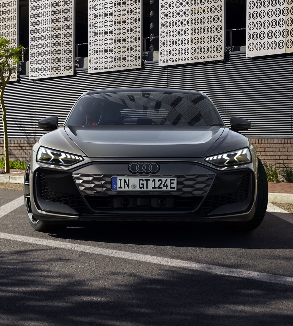 Ein grauer Audi RS e-tron GT steht frontal auf einer Straße mit moderner Metallfassade und Bäumen im Hintergrund