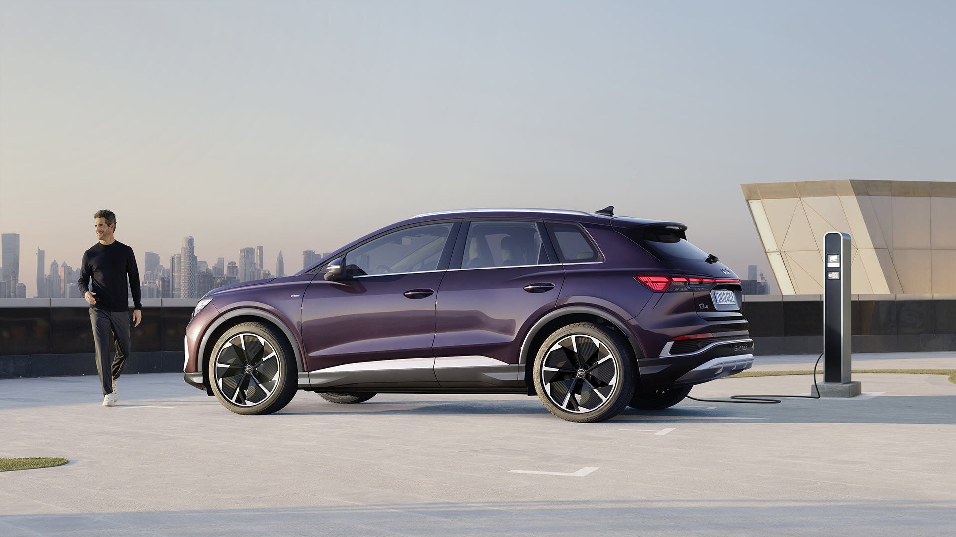 A laughing man approaches an Audi Q4 e-tron that is connected to a charging station on a parking deck with a big city skyline in the background.