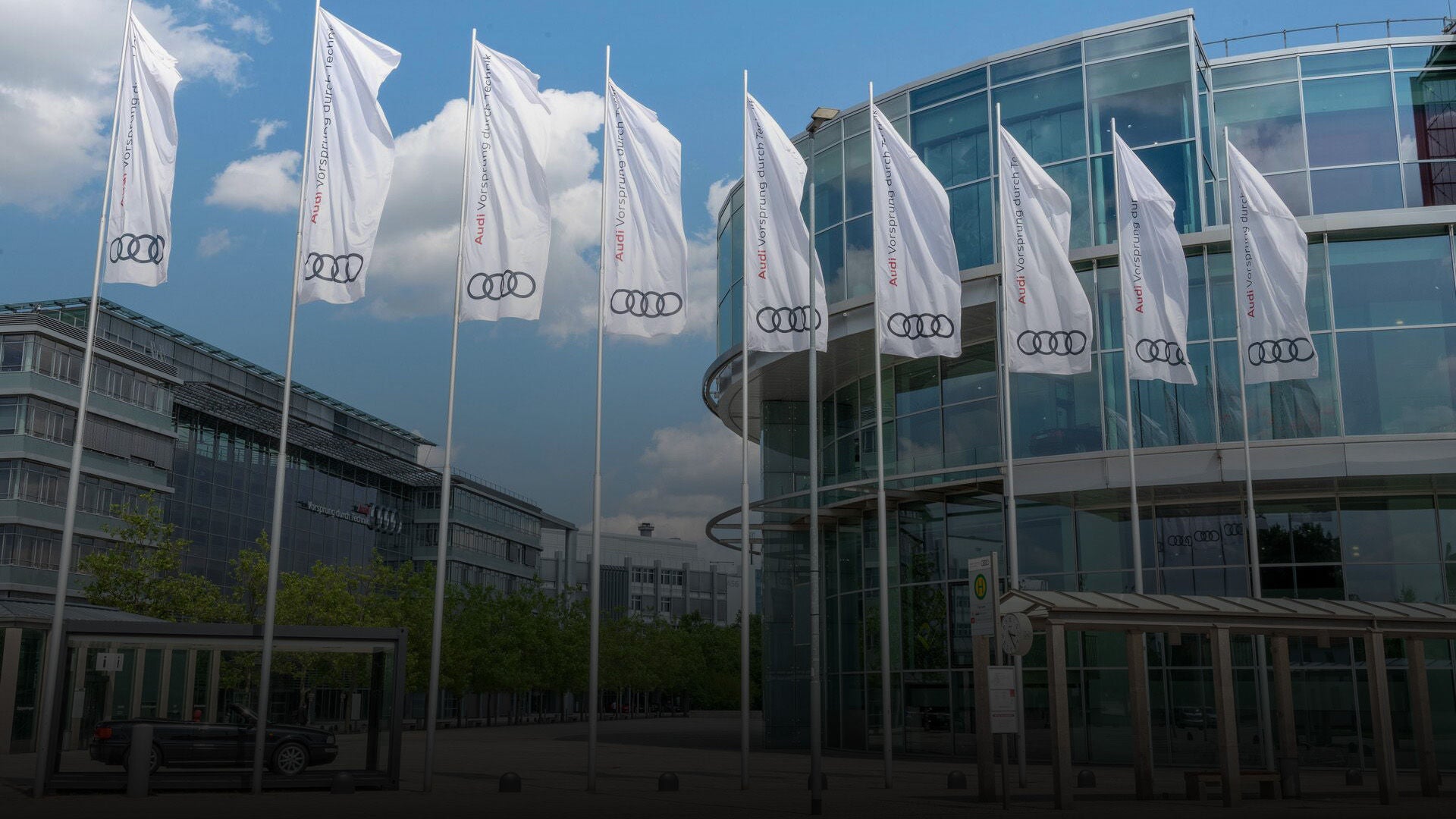 A row of white Audi flags flies in front of a modern glass building under a blue sky.
